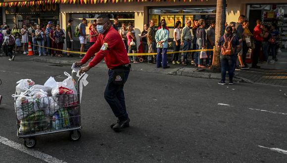 Un empleado de un supermercado empuja un carrito lleno de comestibles mientras las personas hacen cola esperando su turno para ingresar a un supermercado en Medellín, Colombia. (Foto: AFP)