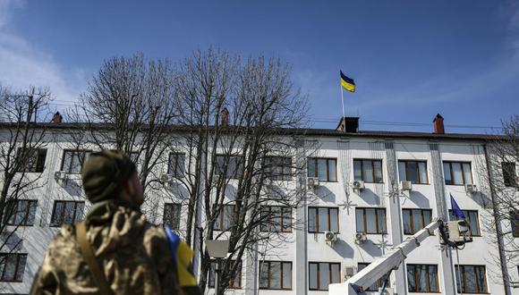 Un militar mira la bandera ucraniana levantada por un empleado en el ayuntamiento de Bucha, en el oeste de Kiev, el 7 de abril de 2022. (Foto de RONALDO SCHEMIDT / AFP)