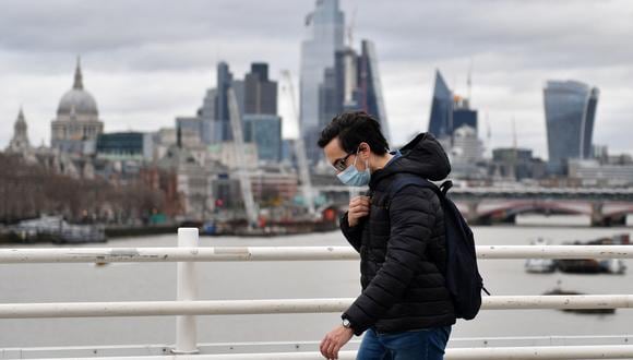 Un peatón que se cubre la cara, debido al covid-19, camina por el puente de Waterloo para cruzar el río Támesis, con rascacielos y oficinas de la ciudad de Londres. (Foto: JUSTIN TALLIS / AFP)