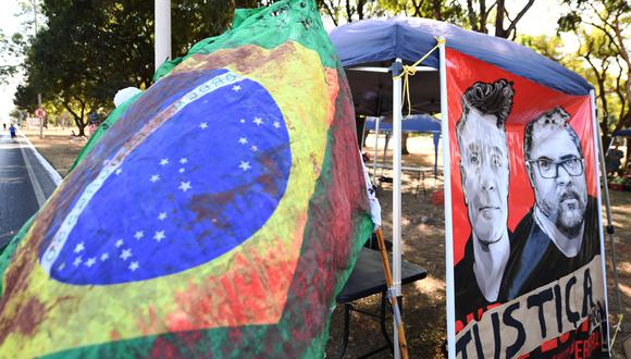 Vista de una bandera brasileña teñida de rojo que representa sangre junto a una pancarta en demanda de justicia por el parentesco del periodista británico Dom Phillips y el indigenista brasileño Bruno Pereira, durante una protesta en Brasilia, el 19 de junio de 2022. (Foto por EVARISTO SA / AFP)