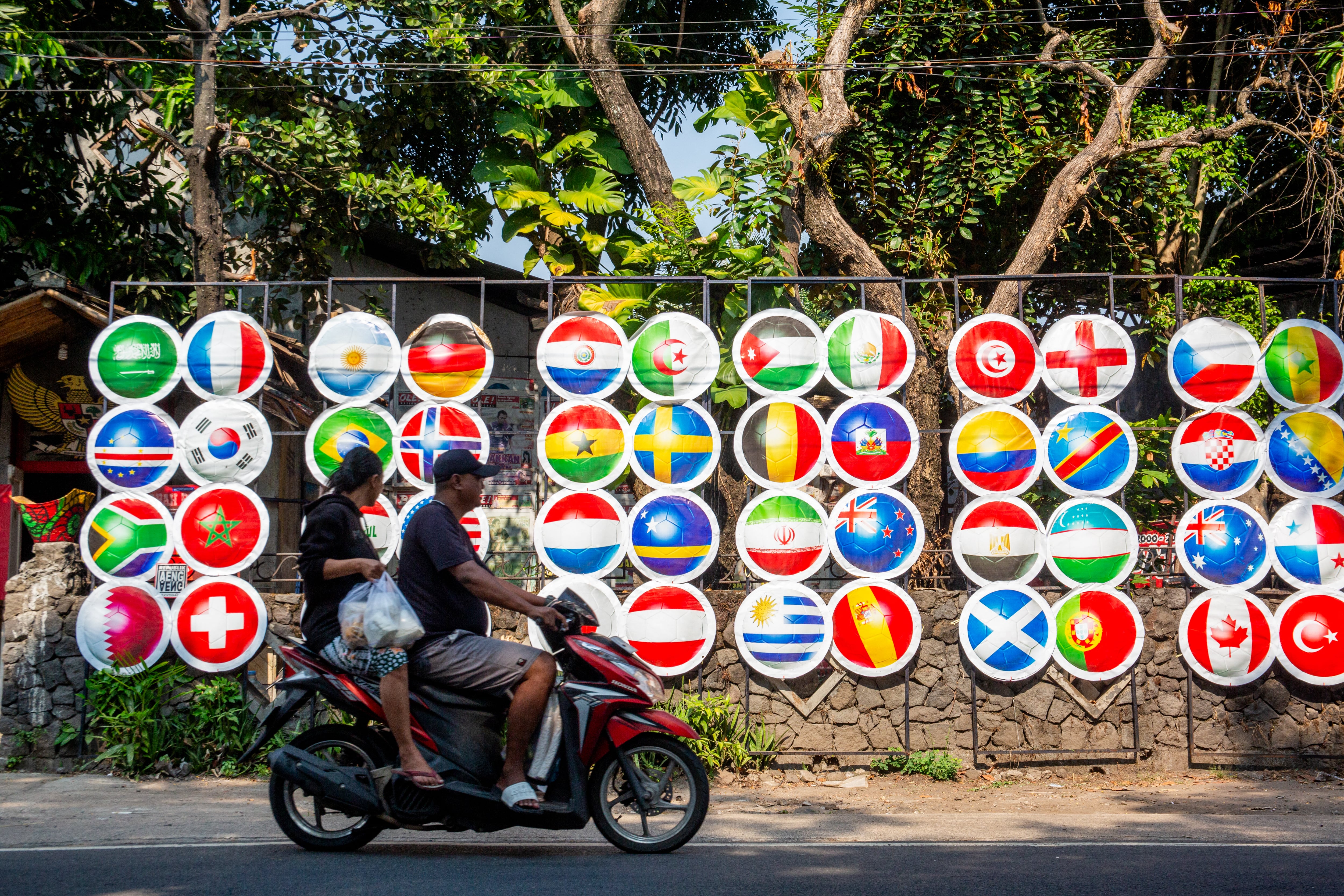 People on a motorcycle ride past decorative ornaments displaying dozens of flags of countries participating in the 2026 FIFA World Cup, set up along a roadside by local residents to celebrate the tournament in Surakarta, Central Java, on July 5, 2026. (Photo by DEVI RAHMAN / AFP)