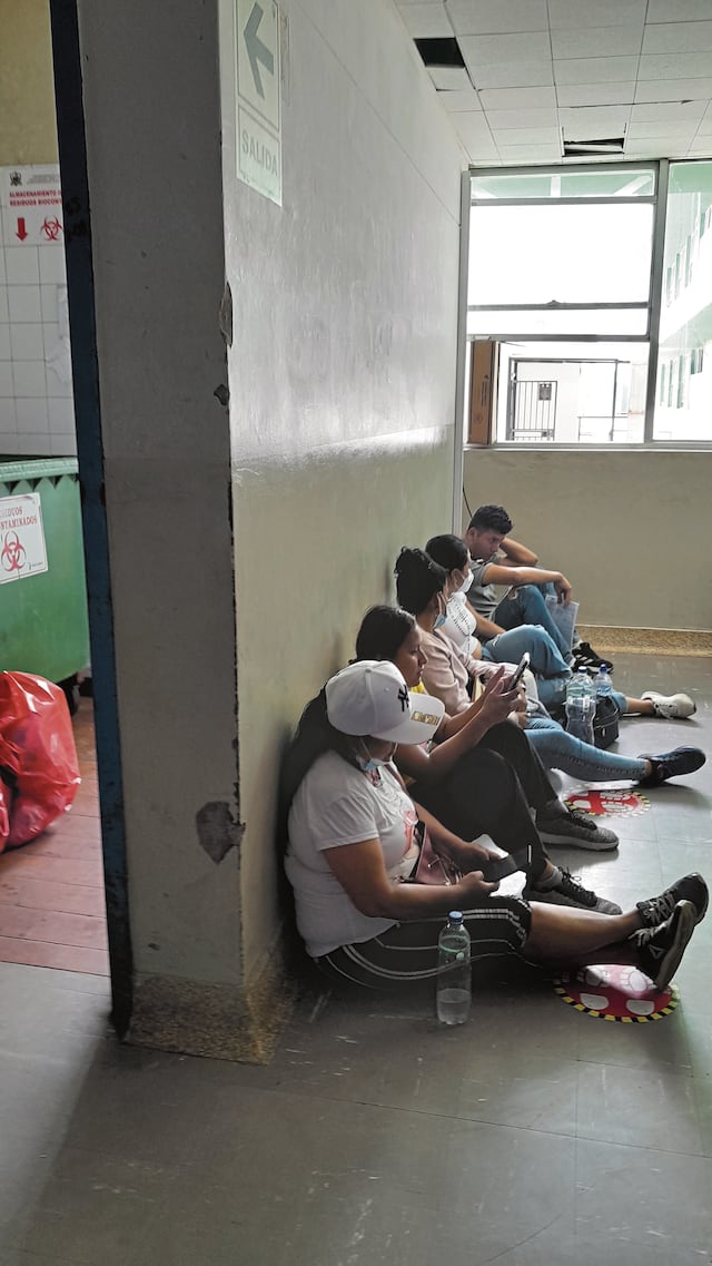 Familiares esperan sentados en el piso. Al costado se observa un cuarto abierto con residuos hospitalarios.